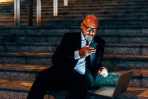 Homme d'affaires mature assis sur les marches avec smartphone et ordinateur portable — Photo de stock