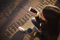 Молода жінка сидить на відкритому повітрі, тримає чашку кави, використовуючи смартфон, татуювання на руках, підвищений вид — стокове фото