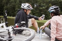 Зрелая пара в велосипедных шлемах смотрит на смартфон на пирсе — стоковое фото