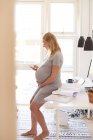 Femme enceinte appuyée contre le bureau et regardant smartphone — Photo de stock