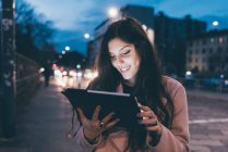Jeune femme, à l'extérieur, la nuit, regardant la tablette numérique, visage illuminé — Photo de stock