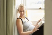 Mujer embarazada con libro sentado por la ventana - foto de stock