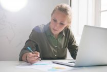 Молода бізнес-леді робить нотатки і використовує ноутбук на офісному столі — стокове фото