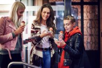 Three young women with ice cream cones looking at smartphones on city street — Stock Photo