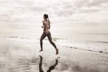 Vista trasera de una joven corredora corriendo descalza a lo largo del borde del agua en la playa - foto de stock