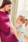 Chica con estetoscopio de juguete escuchando el estómago de la madre embarazada - foto de stock