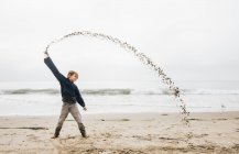 Ritratto di giovane ragazzo sulla spiaggia che lancia sabbia a forma di arco — Foto stock