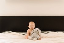 Portrait of cute baby girl sitting on bed with soft toy — Stock Photo