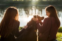 Two young women petting dog on river bank — Stock Photo