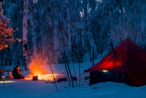 Homem sentado ao lado da fogueira, à noite, perto da tenda, na floresta coberta de neve, Rússia — Fotografia de Stock