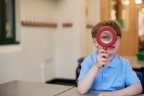 Schüler blickt durch Lupe im Klassenzimmer der Grundschule, Porträt — Stockfoto