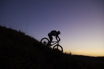 Silhueta de ciclista de montanha masculino montando na colina abaixo ao pôr do sol — Fotografia de Stock