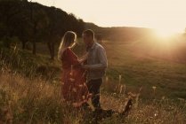 Romantic pregnant couple standing face to face on hillside at sunset — Stock Photo