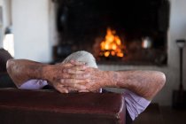 Rear view of senior man at home with hands behind his head in front of log fire — Stock Photo