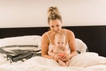 Baby girl and mother sitting in bed and looking at smartphone — Stock Photo