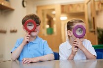 Schoolboy and girl looking through magnifying glasses in classroom at primary school, portrait — Stock Photo