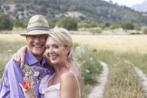 Couple dans le champ avec un bouquet de fleurs étreignant, regardant la caméra souriant — Photo de stock