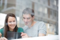 Hombre y mujer adultos en la cafetería, mirando la tableta digital, vista a través de la ventana - foto de stock