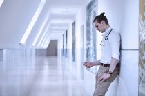 Doctor in hospital corridor leaning against wall using smartphone — Stock Photo