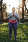 Викривлена молода жінка тренується в парку і дивиться на смартфон — стокове фото