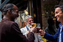 Tre uomini maturi, in piedi fuori pub, tenendo bicchieri di birra, facendo un brindisi — Foto stock