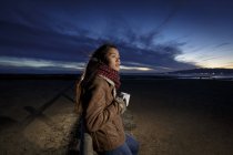 Junge Frau in Schal gehüllt schaut in der Abenddämmerung vom Strand aus — Stockfoto