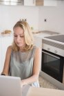 Junge Frau tippt auf Laptop am Küchentisch — Stockfoto
