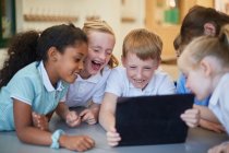 Schoolboys and girls laughing at digital tablet in classroom at primary school — Stock Photo