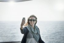 Mulher de óculos de sol tomando selfie com mar no fundo — Fotografia de Stock