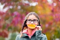Retrato de menina com tranças e óculos cobrindo a boca com folha olhando para longe — Fotografia de Stock