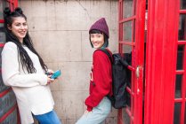 Portrait of two young stylish women leaning against red phone boxes, London, UK — Stock Photo
