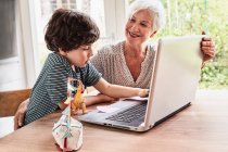 Abuela y nieto sentado en la mesa, utilizando el ordenador portátil - foto de stock