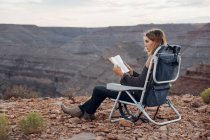 Junge Frau sitzt auf Campingstuhl und liest Buch, mexikanischer Hut, utah, usa — Stockfoto