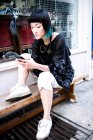 Молода стильна жінка сидить за межами магазину зі смартфоном — стокове фото