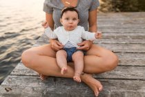 Mãe sentada cruz pernas no lago cais com bebê filha no colo, cortado — Fotografia de Stock