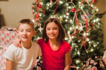 Портрет брата и сестры, улыбающихся в камеру на фоне рождественской елки — стоковое фото