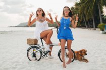 Portrait of two young women with bicycle making peace sign on sandy beach, Krabi, Thailand — Stock Photo