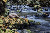 Vista lateral de mujer desnuda haciendo yoga sobre roca por río - foto de stock