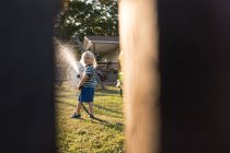 View through gap in fence of boy spraying water from hosepipe — Stock Photo