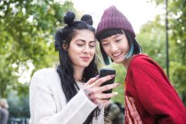 Дві молоді стильні жінки дивляться на смартфон у міському парку — стокове фото