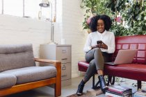 Женщина в офисе сидит на диване с ноутбуком, пишет смс на смартфоне — стоковое фото