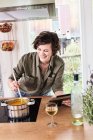 Mujer adulta media removiendo la olla en la cocina, sosteniendo la tableta digital, riendo - foto de stock