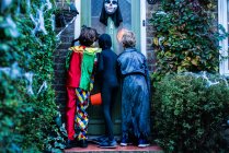 Tres niños en disfraces de Halloween, de pie en la puerta, truco o trato, vista trasera - foto de stock