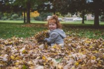 Portrait of red haired female toddler in park with bundles of autumn leaves — Stock Photo