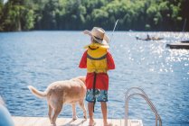 Вид сзади на собаку и мальчика в ковбойской шляпе, рыбалка с причала озера — стоковое фото