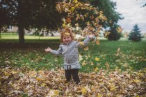 Portrait of red haired female toddler in park throwing autumn leaves — Stock Photo