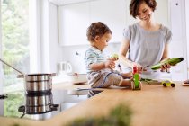 Baby-Mädchen sitzt auf Küchentisch, während Mutter Gurke schneidet — Stockfoto