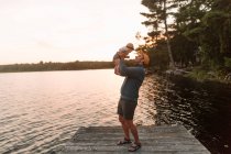 Man holding up baby daughter on lake pier — Stock Photo