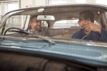 Дві машини механіки розмовляють на передньому сидінні старовинного автомобіля в ремонтному гаражі — стокове фото