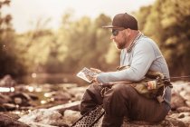 Рыбак, сидящий на речных скалах, глядя на смартфон, Мозирье, Брезовица, Словения — стоковое фото